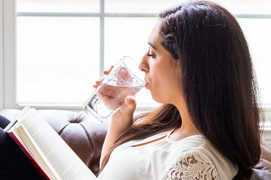 Young woman drinking water while sitting on sofa reading book